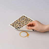 Hand holding a leopard print coaster over a coffee spill on a gray surface